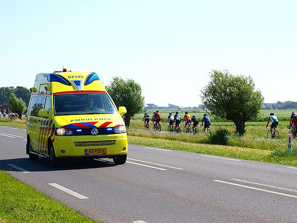 Terwijl een ambulance, met aan boord een zwaargewonde motorrijdster, met grote spoed over de N354 bij Woudsend naar het ziekenhuis van Sneek rijdt, passeren op de achtergrond deelnemers aan de fietselfstedentocht.