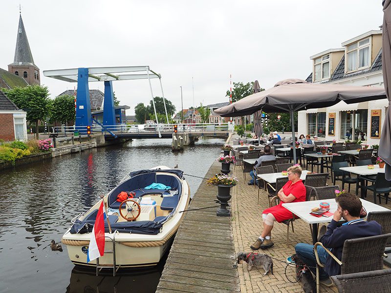 Sloepvaarders uit Earnewâld willen naar de historische binnenstad van Leeuwarden, maar stuiten op de lage ophaalbrug in Warten die twee keer midden op de dag een uur dicht is. Sommigen wachten op het terras van De Kok en de Walvis op wat gebeuren gaat: niks.